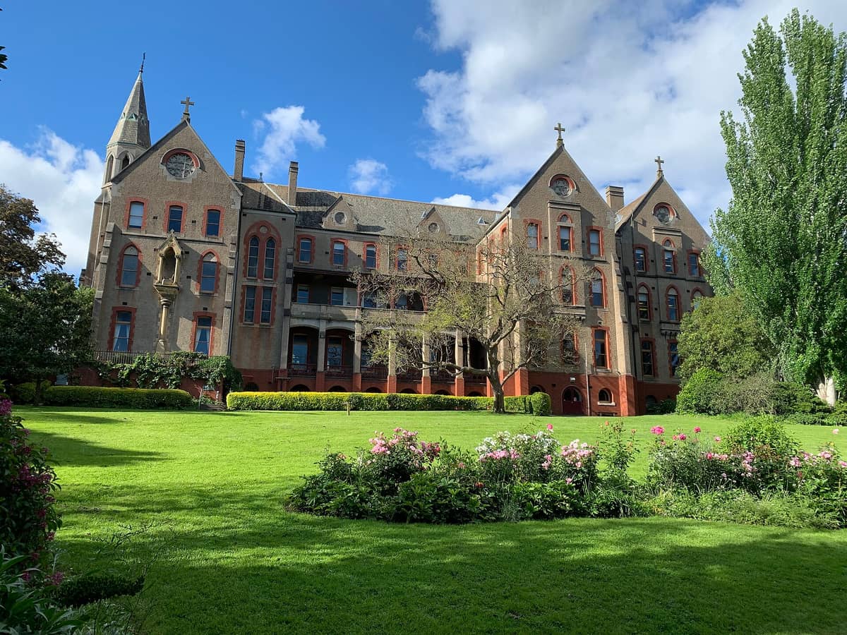 Weitläufige Ansicht des Abbotsford Convent mit seinem roten Backstein- und Steingebäude, Kreuzen auf den steilen Giebeln, saftig grünem Rasen und blühenden Blumenbeeten unter strahlend blauem Himmel.