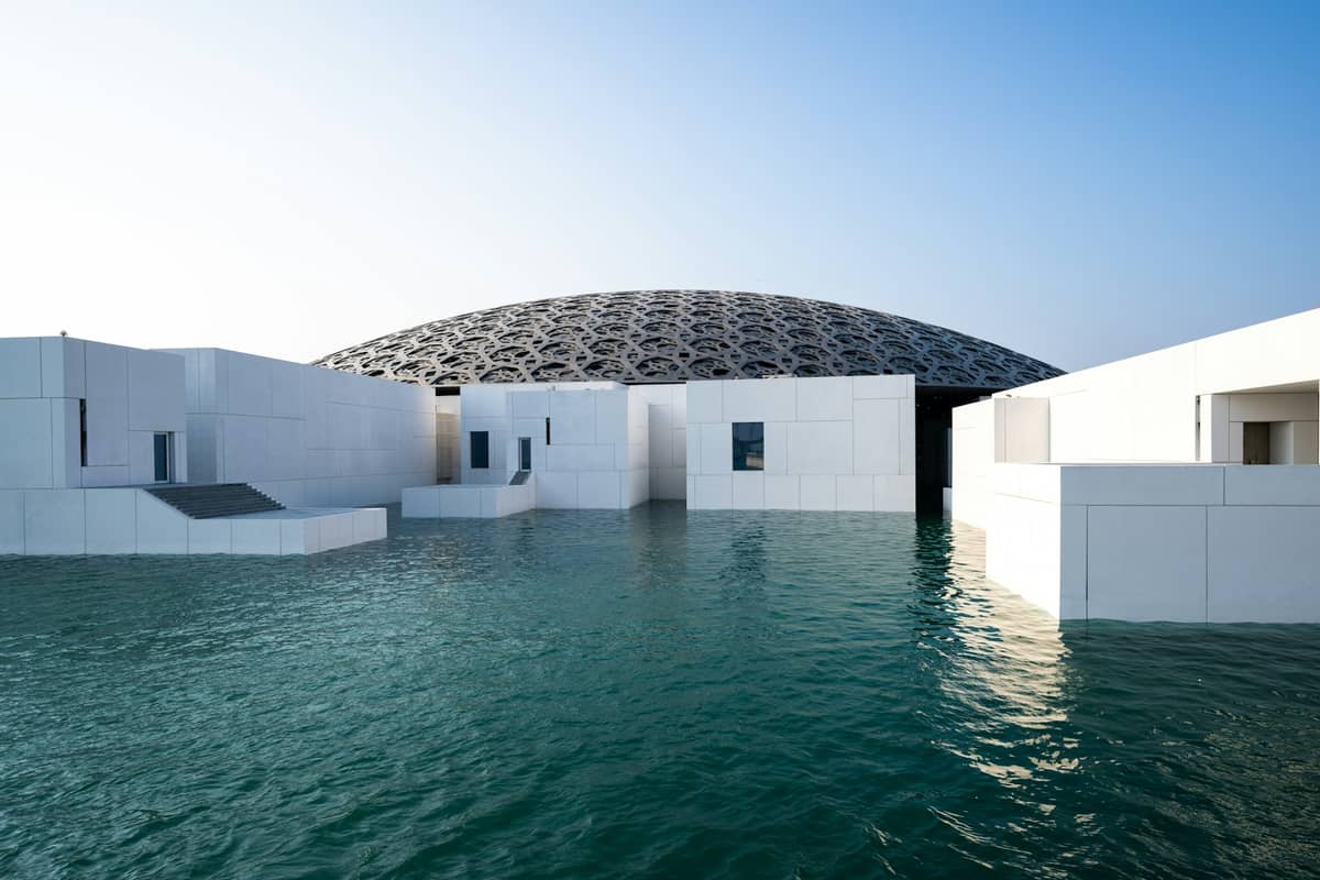 Landschaftliche Außenansicht des Louvre Abu Dhabi mit seiner ikonischen Kuppel und weißen modernen Gebäuden, umgeben von türkisfarbenem Wasser.