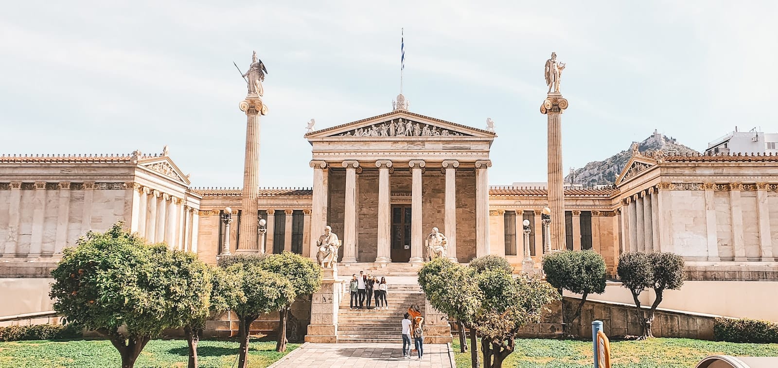 Vista panoramica dell'Accademia di Atene con colonne neoclassiche, statue e vegetazione rigogliosa, che mostra la straordinaria architettura in una giornata luminosa e serena.