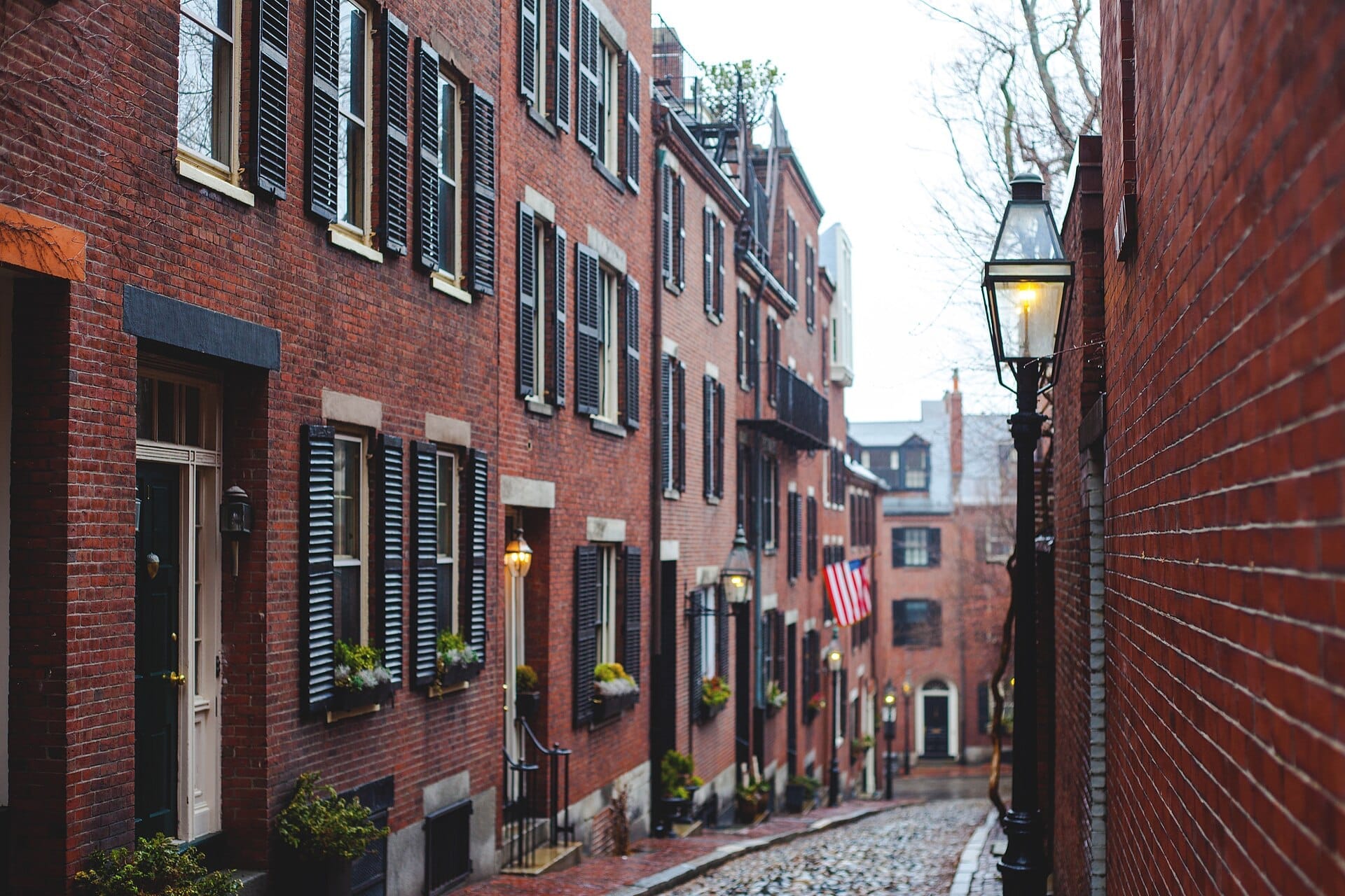 Case a schiera in mattoni rossi con persiane nere fiancheggiano il selciato di Acorn Street a Boston, con lampioni a gas che brillano sotto una luce diurna coperta.
