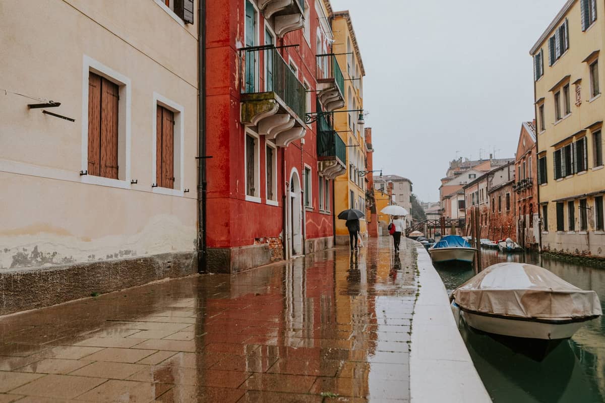 Venezianischer Kanal mit Acqua alta, Spiegelungen auf nassen Gehwegen, bunte Gebäude, Menschen mit Schirmen auf überschwemmten Wegen.