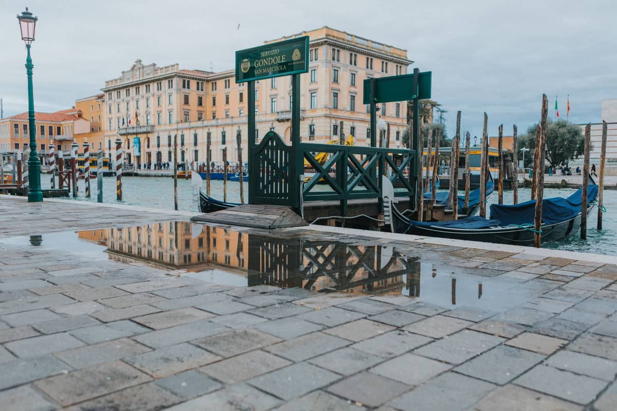 Venezianische Uferpromenade mit einer kleinen Pfütze vom Hochwasser auf einem Steinweg, Gondelsteg und klassischen Gebäuden im Hintergrund.