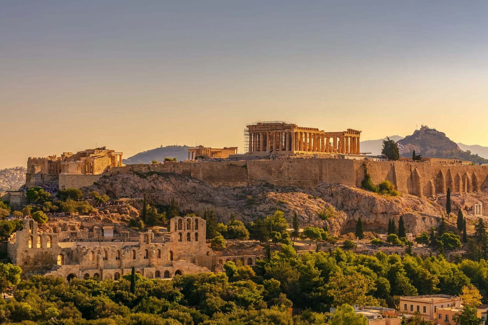 Vista dell'Acropoli di Atene al tramonto, con il Partenone e le rovine antiche circostanti sopra una lussureggiante vegetazione e il paesaggio urbano.