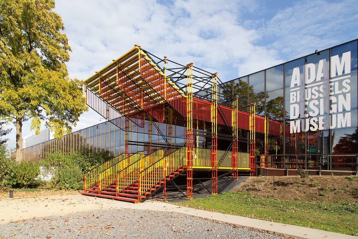 Exterior do Design Museum Brussels com grandes painéis de vidro espelhado, letras brancas em negrito e uma colorida instalação de escadaria exterior em amarelo e vermelho.