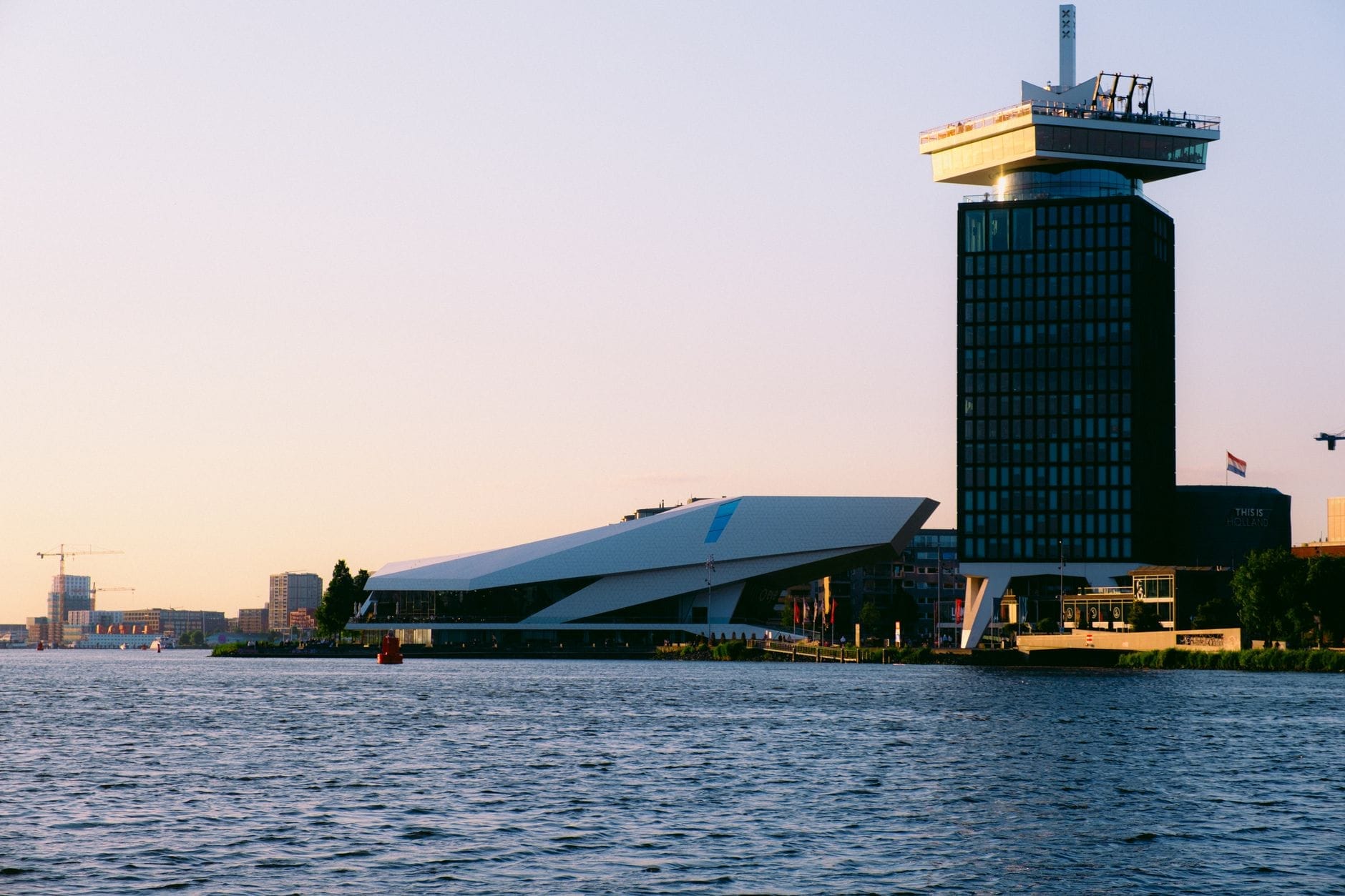 La torre A'DAM Lookout y el EYE Filmmuseum al atardecer, vistos desde el otro lado del río IJ, con el agua en calma y una cálida luz dorada.