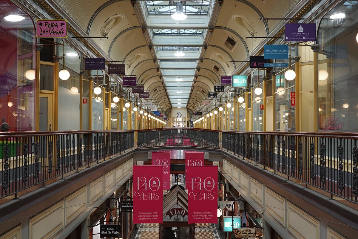Interior of Adelaide Arcade showing Victorian architecture, glass roof, ornate railings, and specialty shops with signs and banners celebrating 140 years.