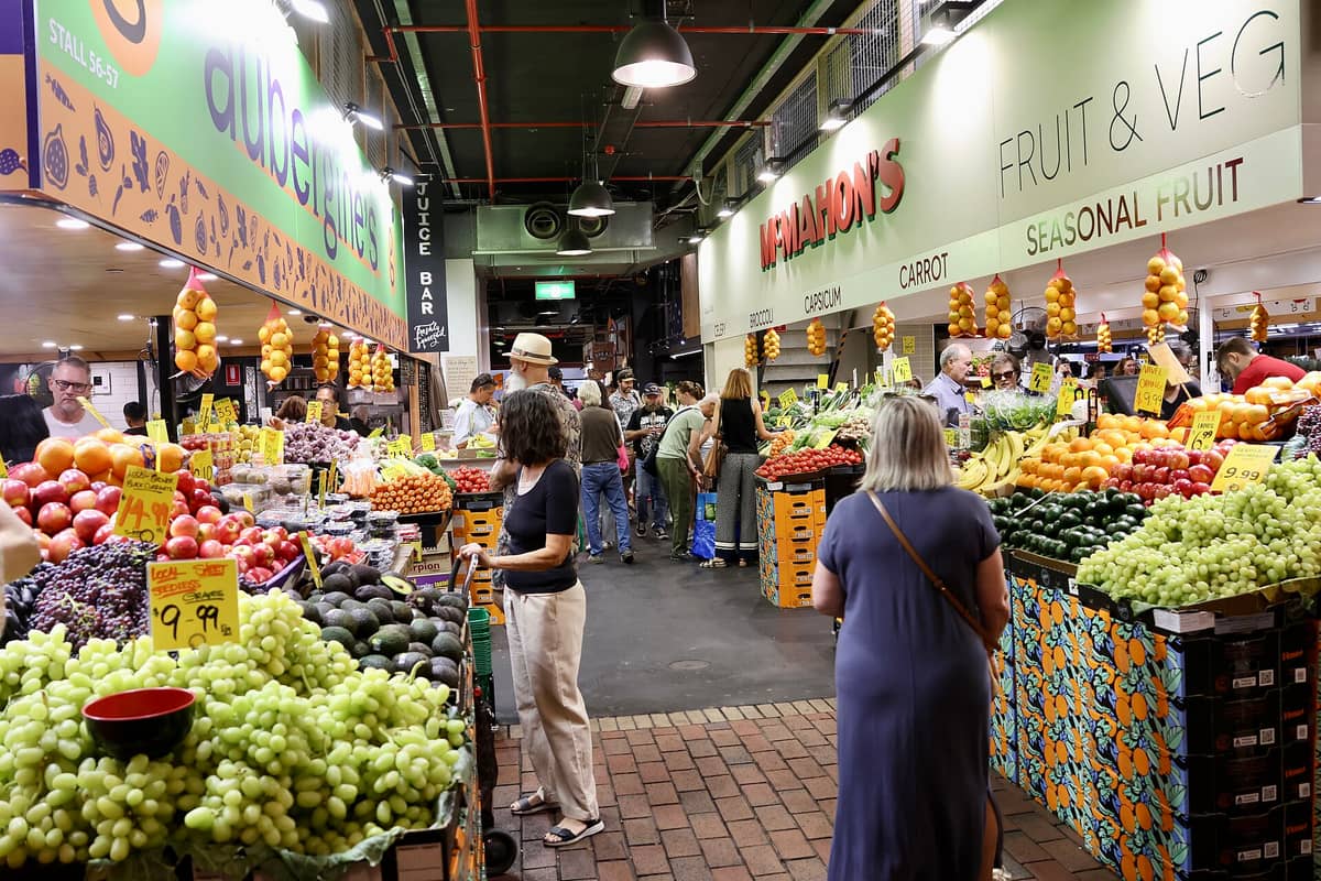 Wide interior view of Adelaide Central Market showing fruit and vegetable stalls, shoppers, colorful produce, and signage for McMahon's and aubergine's