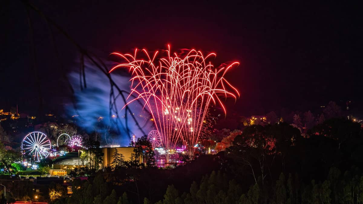 Czerwone fajerwerki wybuchają nad rozświetlonym terenem festiwalowym z diabelskimi młynami i karuzelami w nocy, otoczonym drzewami w Adelajdzie.