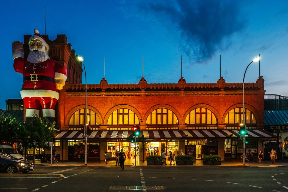 Widok uliczny budynku Adelaide Central Market o zmierzchu z podświetlonymi witrynami sklepów i dużym nadmuchiwanym Mikołajem na dachu.