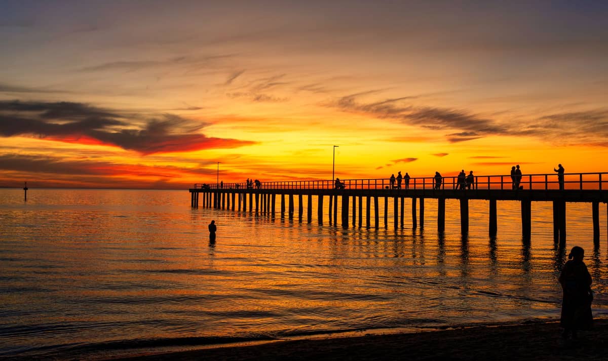 Silhouetten von Menschen auf einem langen Pier bei Sonnenuntergang mit leuchtendem orangefarbenem Himmel, der sich im ruhigen Ozean spiegelt, klassische Küstenszene von Adelaide.
