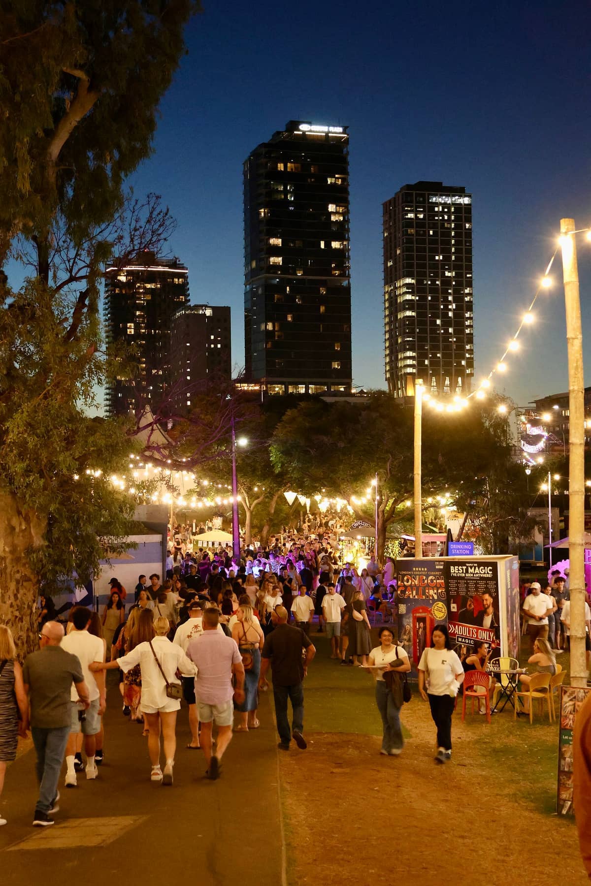 Multitudes de personas pasean bajo las luces del festival por la noche durante el Adelaide Fringe, con altos edificios de la ciudad iluminados al fondo.