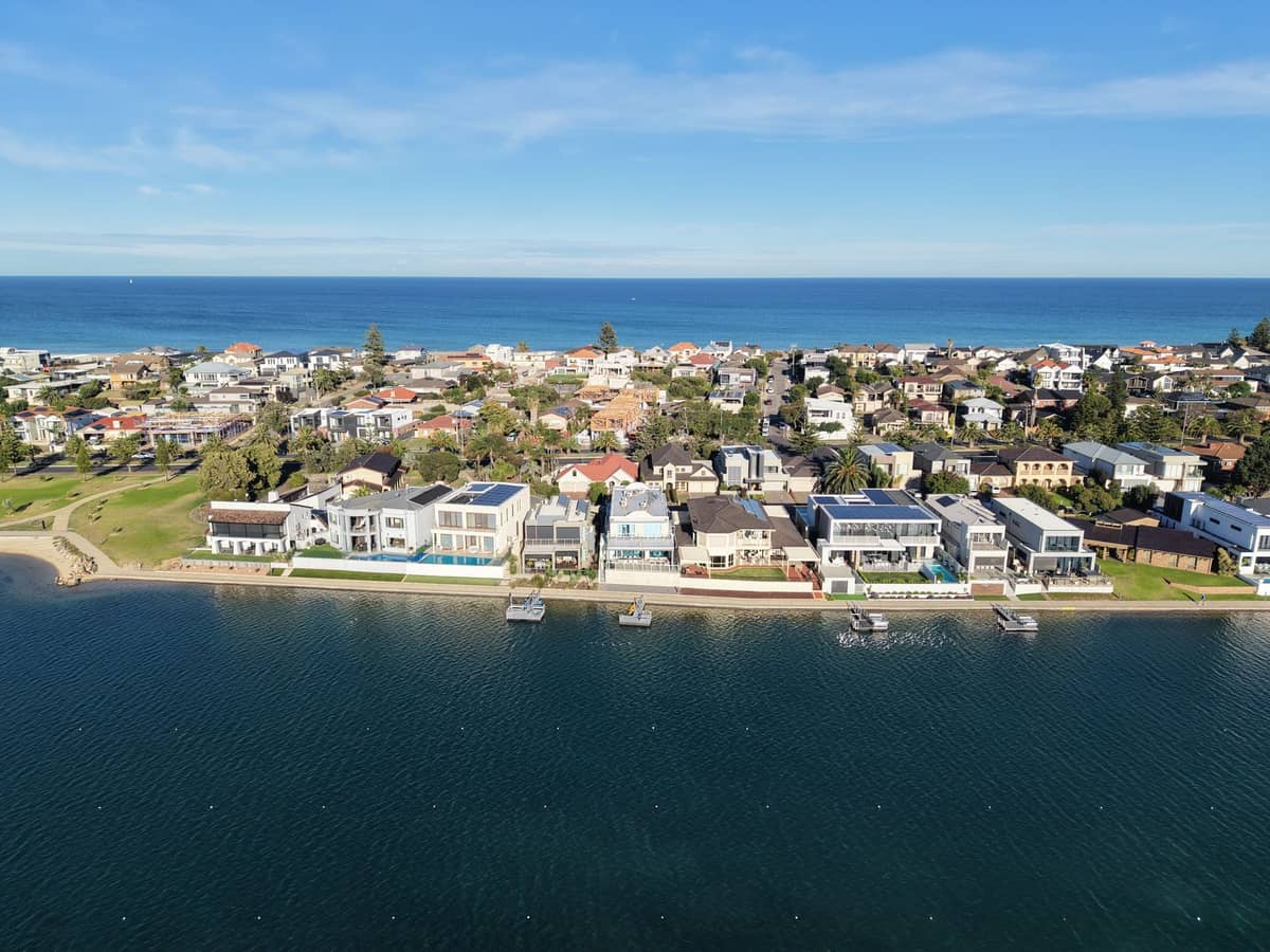 Aerial view of luxury waterfront homes in an upscale Adelaide neighbourhood, with clear blue water and ocean in the background on a sunny day.