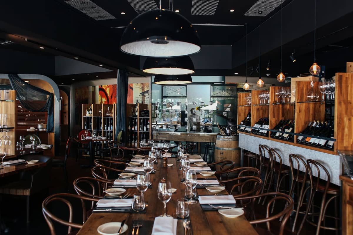Elegant, upscale restaurant with long wooden table, stylish lighting, wine shelves, and place settings ready for fine dining.
