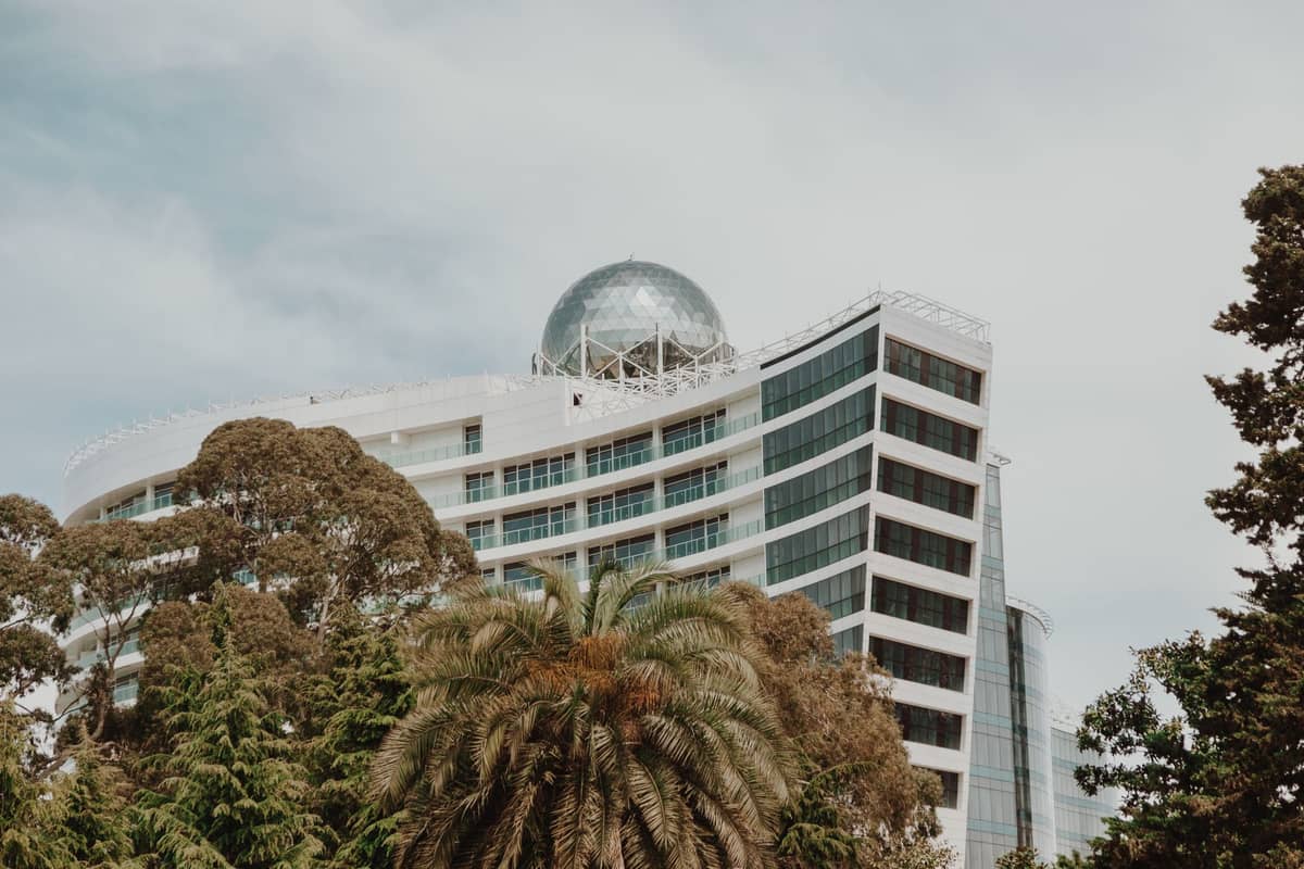 Modern curved hotel building with glass facade and large dome structure, surrounded by palm trees and greenery in Adelaide.