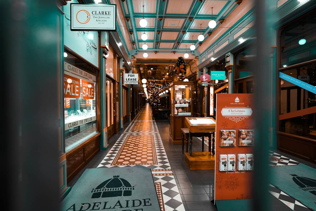 Nocny widok wnętrza Adelaide Arcade z wycieraczką z logo Adelajdy, witrynami sklepowymi, ciepłym oświetleniem i zdobnym sufitem — klimat miejskiego nocnego życia.