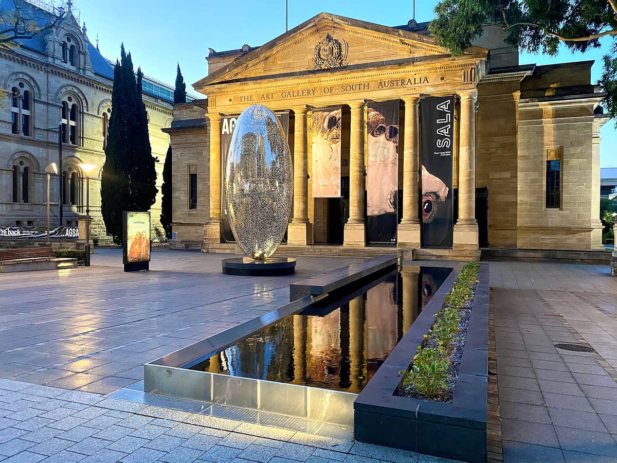 Esterno dell'Art Gallery of South Australia con striscioni e una grande scultura riflettente davanti, alla luce della sera.