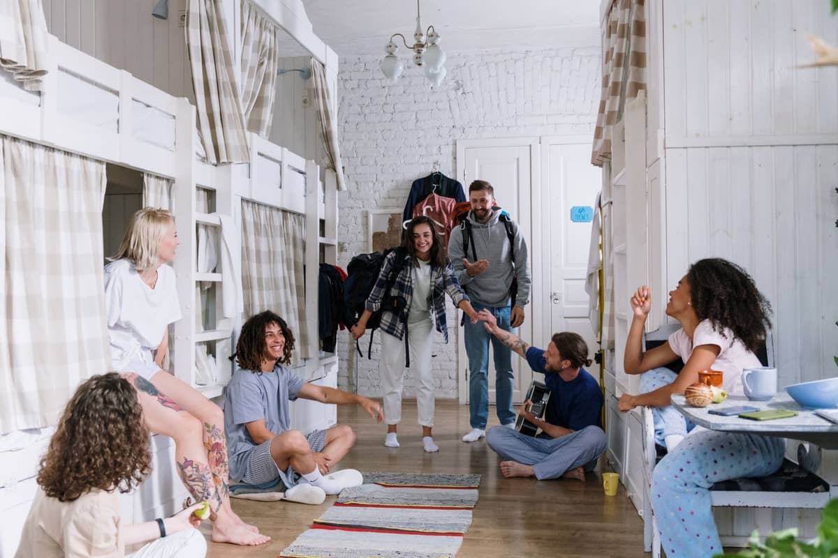 Giovani viaggiatori seduti e in conversazione in una camera dormitorio di un ostello moderno e pulito con letti a castello bianchi e un'atmosfera rilassata e sociale.