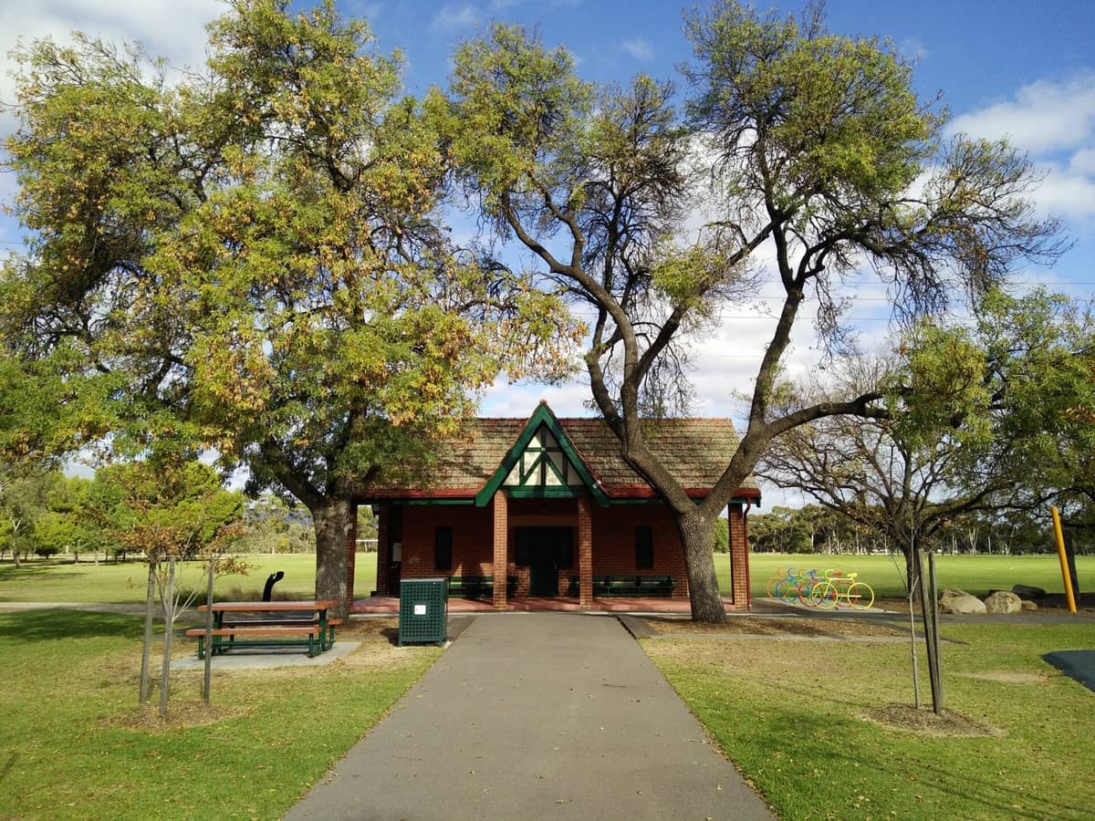 A red-brick pavilion surrounded by leafy green trees and open lawns in Adelaide Park Lands under a bright blue, partly cloudy sky.