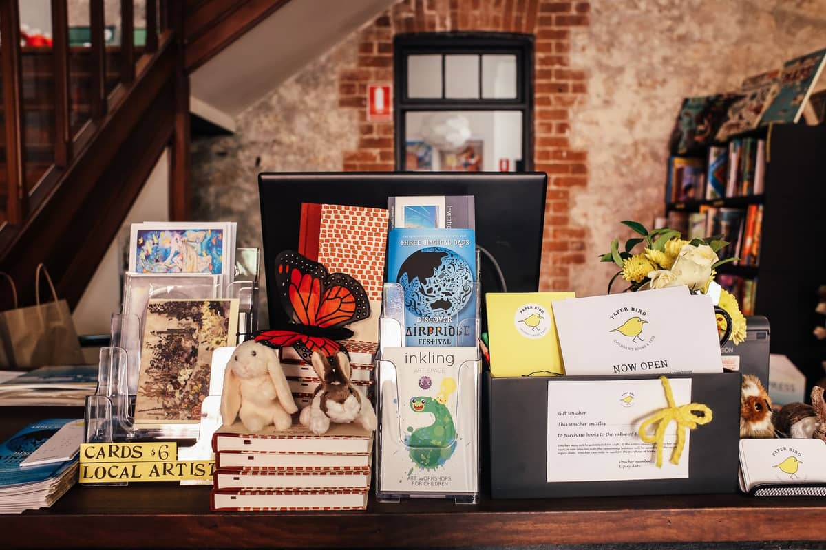 Counter with cards, books, and a sign for local arts gift shop, with brick and stone wall in the background.