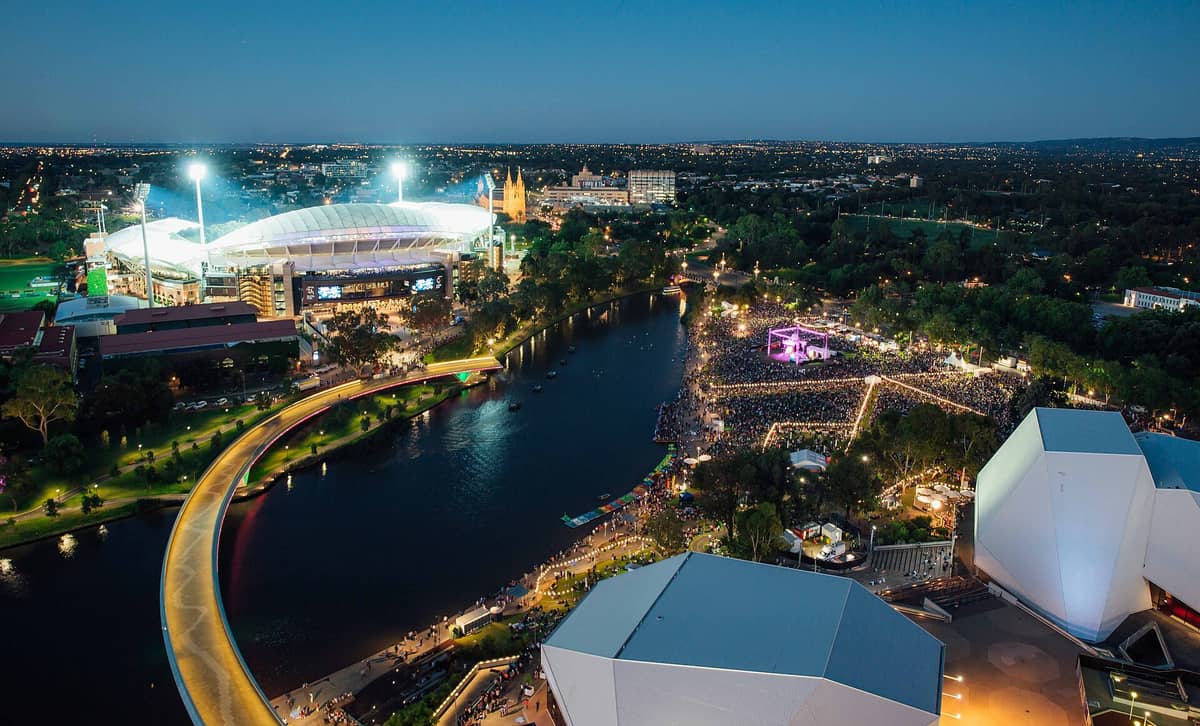 Aerial night view of Adelaide Festival Centre, Adelaide Oval, River Torrens, and large festival crowd in the West End arts precinct.