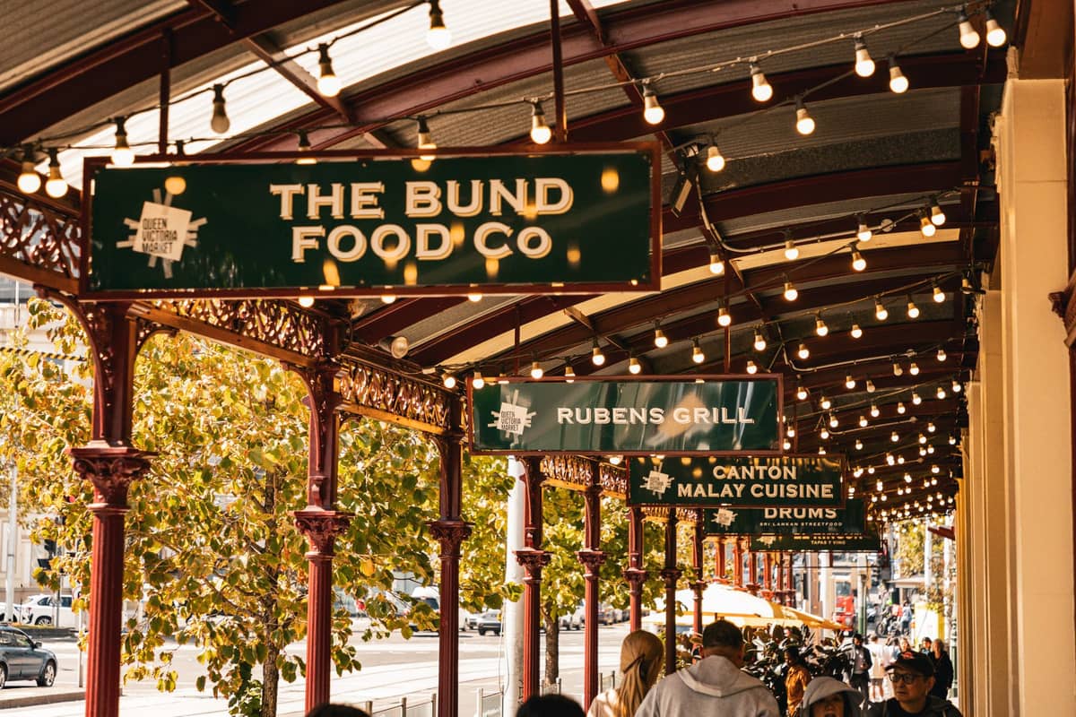 Outdoor arcade at Adelaide Central Market with food vendor signs, string lights, and people walking beneath the canopy in Adelaide’s West End.