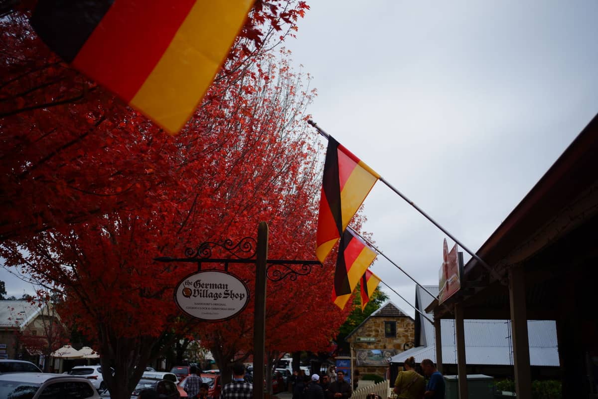 Straßenszene im Dorf Hahndorf mit deutschen Flaggen, roten Herbstbäumen, Fußgängern, Autos und Schildern lokaler Geschäfte.