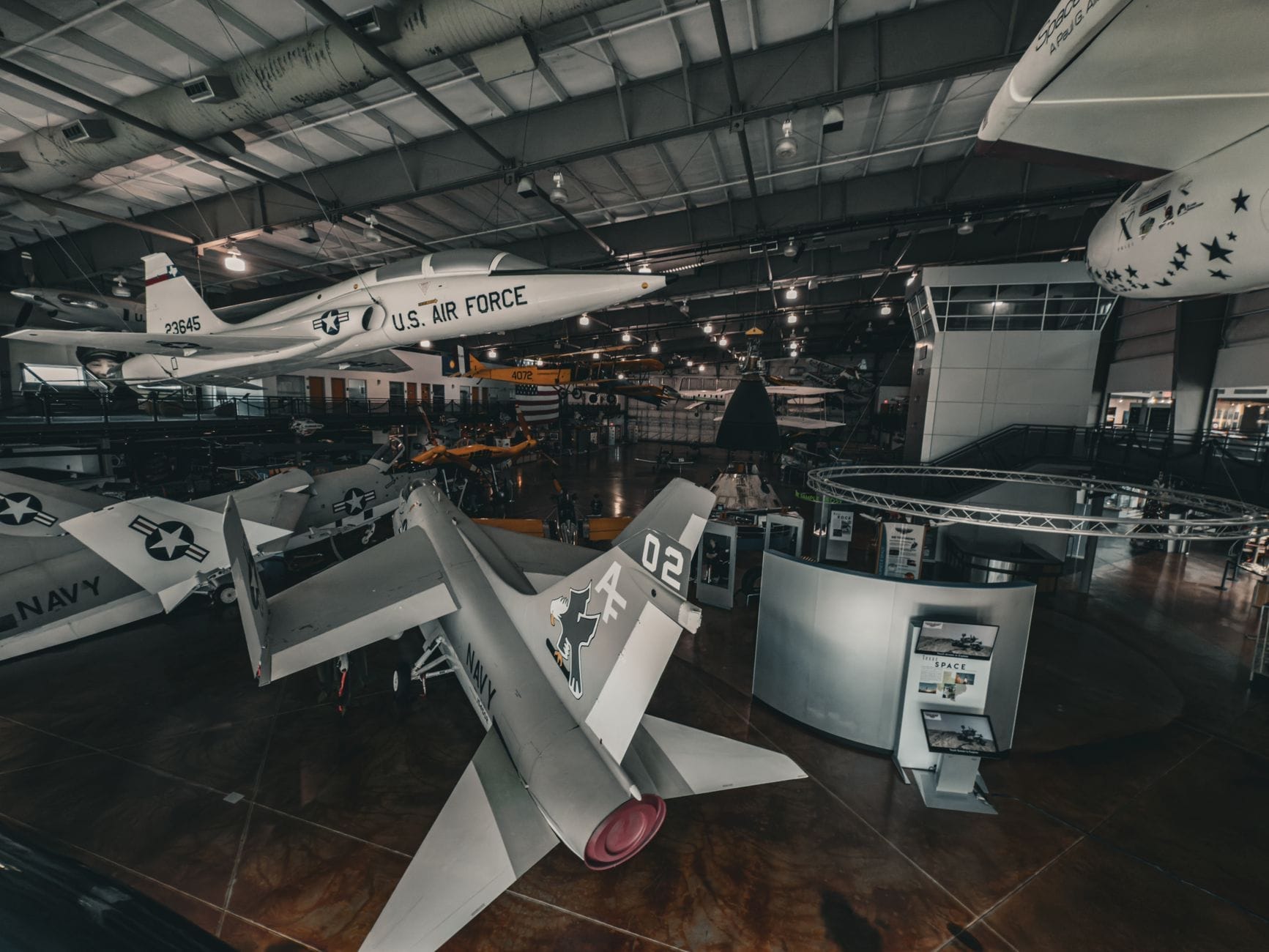 Vista interior de una exhibición de museo de aviación con varios aviones históricos, incluido un jet de la Fuerza Aérea de los EE. UU., bajo un gran techo metálico.