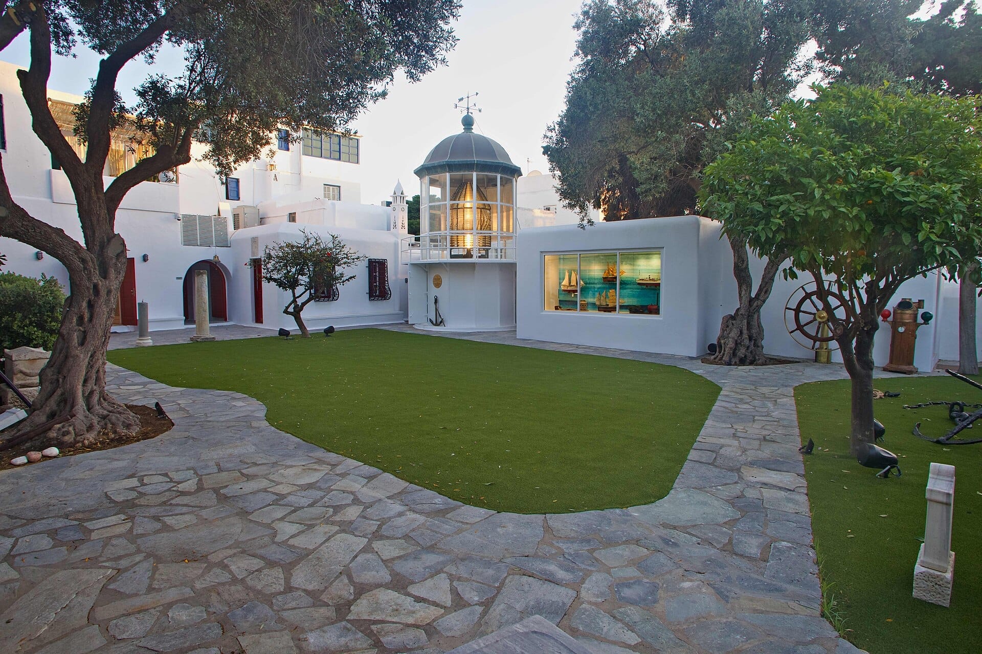 Patio del Museo Marítimo del Egeo en Mykonos, con edificios cicládicos blancos, césped verde, árboles y una ventana con maquetas de barcos.