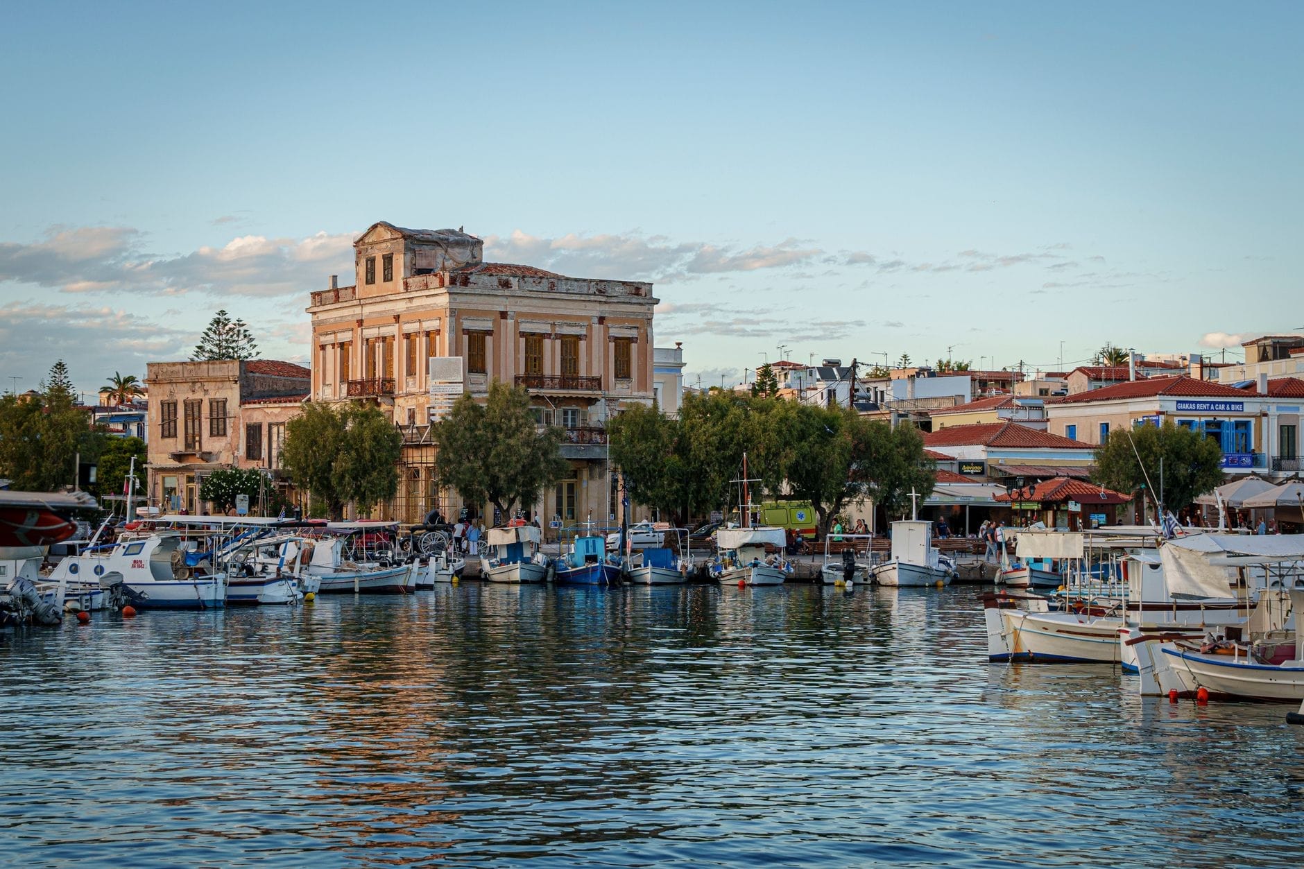 Il caratteristico lungomare di Egina con i suoi edifici tradizionali, le piccole barche ormeggiate nel vivace porto e i riflessi nitidi sull'acqua sotto un cielo azzurro intenso.