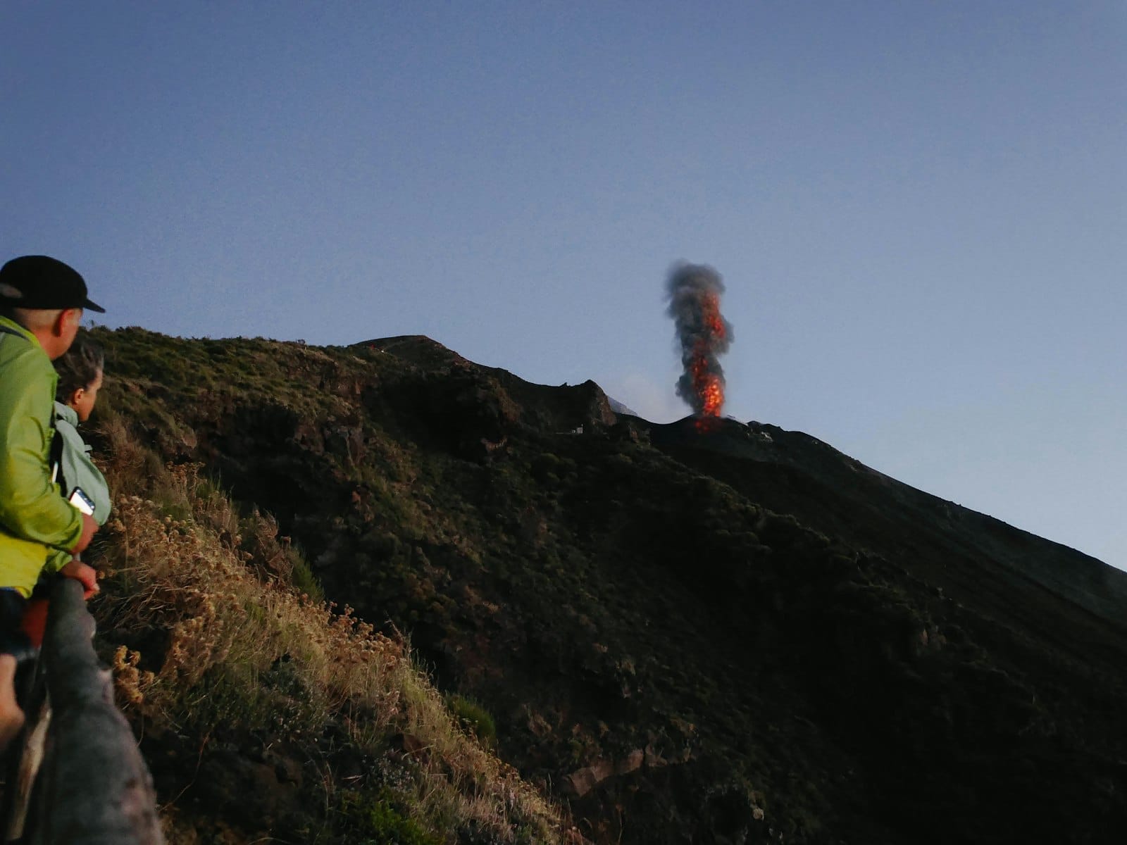 Zwei Personen beobachten den ausbrechenden Vulkan Stromboli in der Dämmerung an einem felsigen grünen Hang auf den Äolischen Inseln, mit sichtbarem Rauch und Lava.