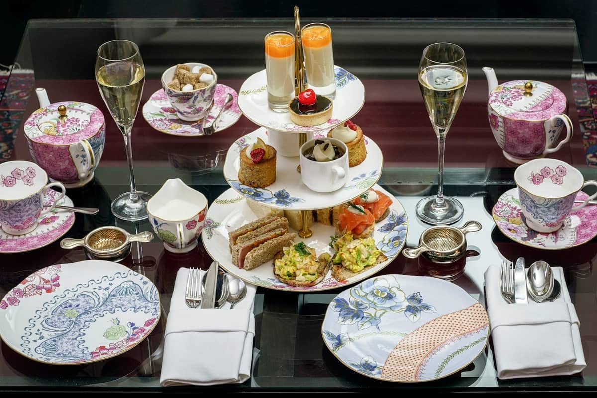 Elegante serviço de chá da tarde com bandeja em camadas de sanduíches, doces e scones, xícaras de chá florais e taças de champanhe sobre uma mesa.