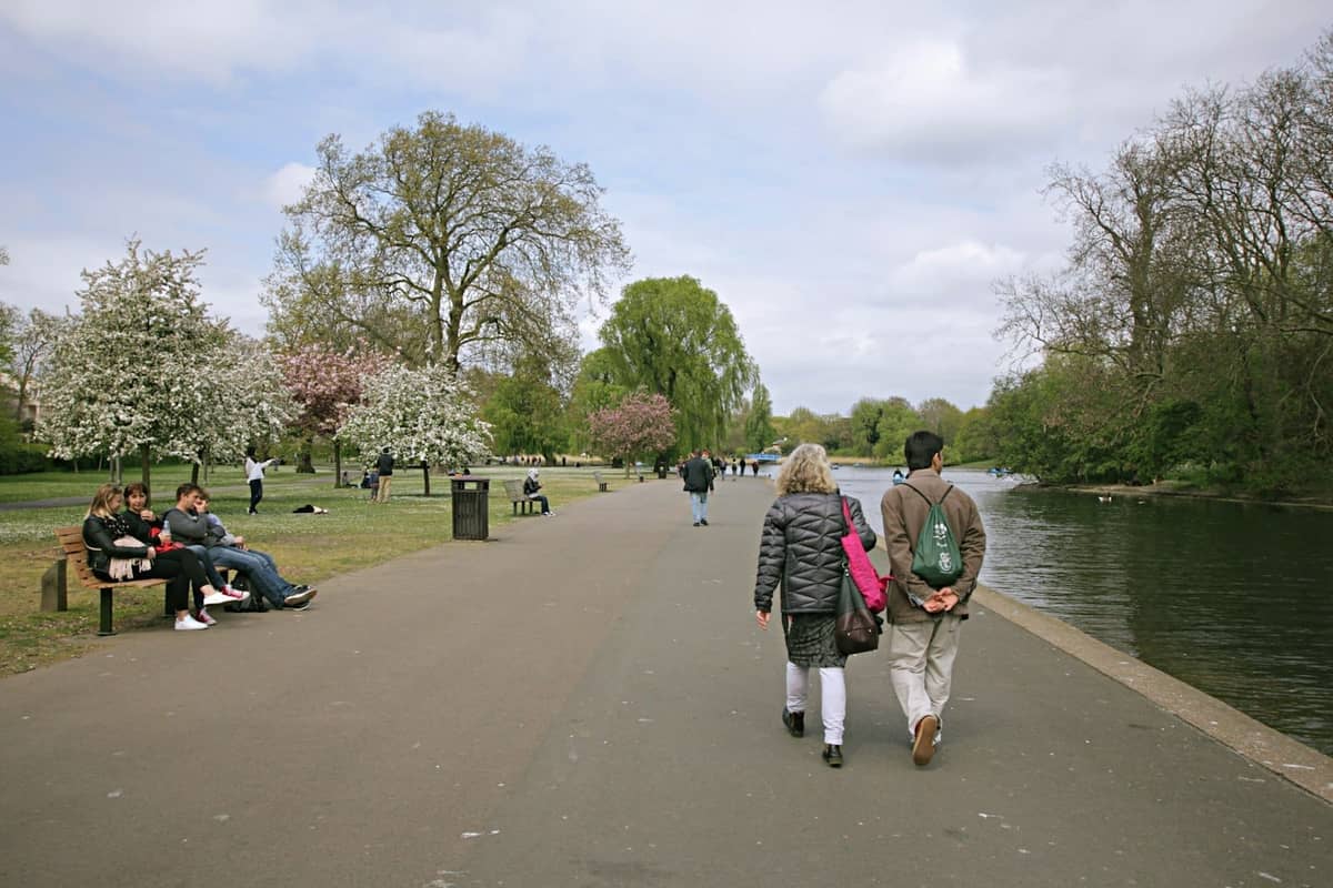 Pessoas caminhando e relaxando à beira do lago em um grande parque de Londres com flores de primavera e caminhos abertos.