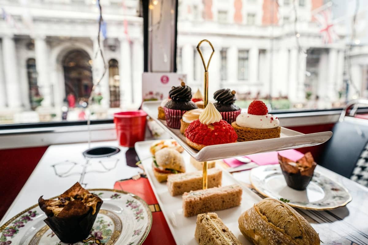 Suporte de três andares com scones, sanduíches de dedo e doces sobre uma mesa junto a uma janela com vista para a arquitetura clássica de Londres.