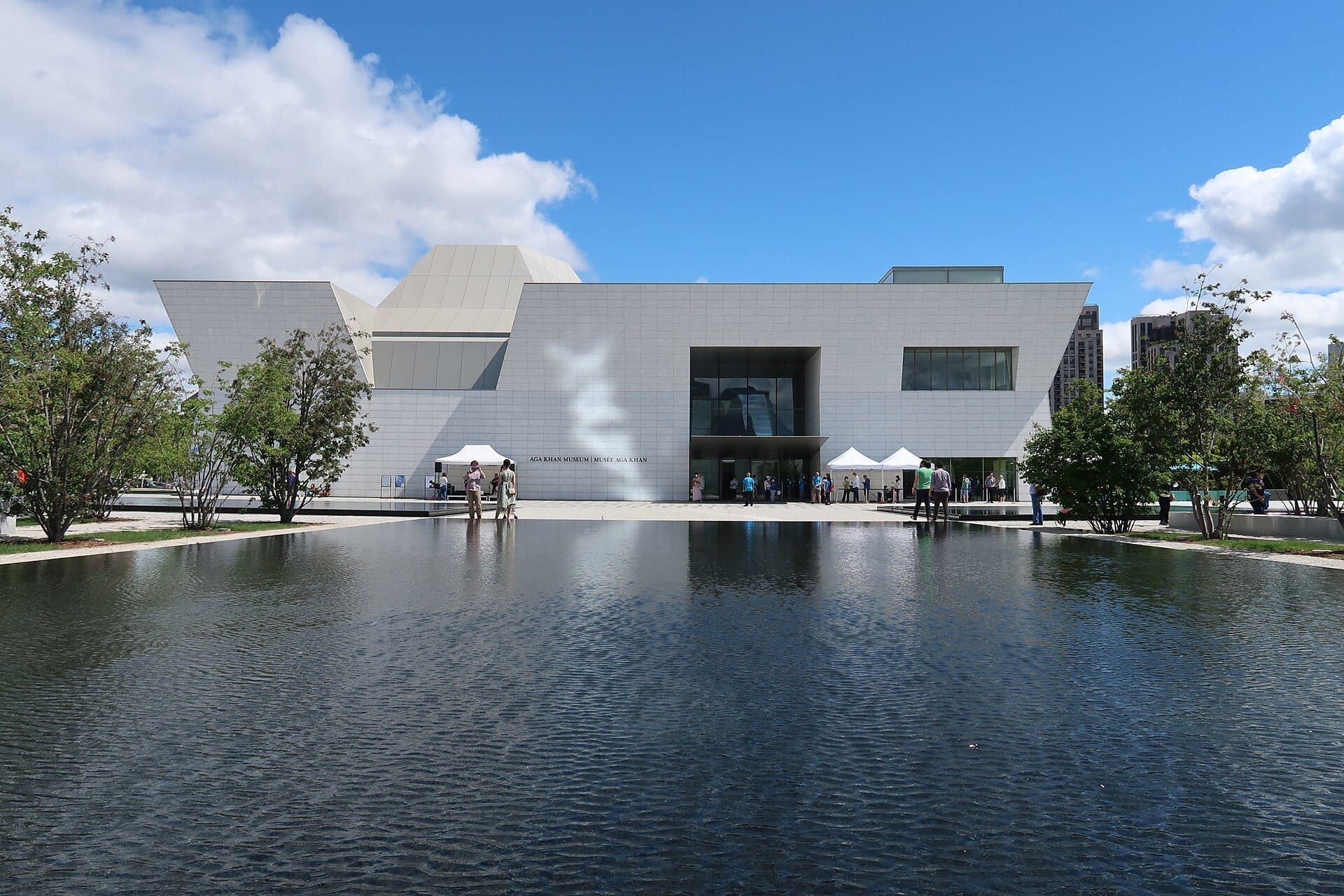 อาคารสีขาวทันสมัยของ Aga Khan Museum ในเมือง Toronto มองเห็นผ่านสระน้ำสะท้อนแสงพร้อมผู้เยี่ยมชมด้านนอกใต้ท้องฟ้าสีฟ้าสดใส