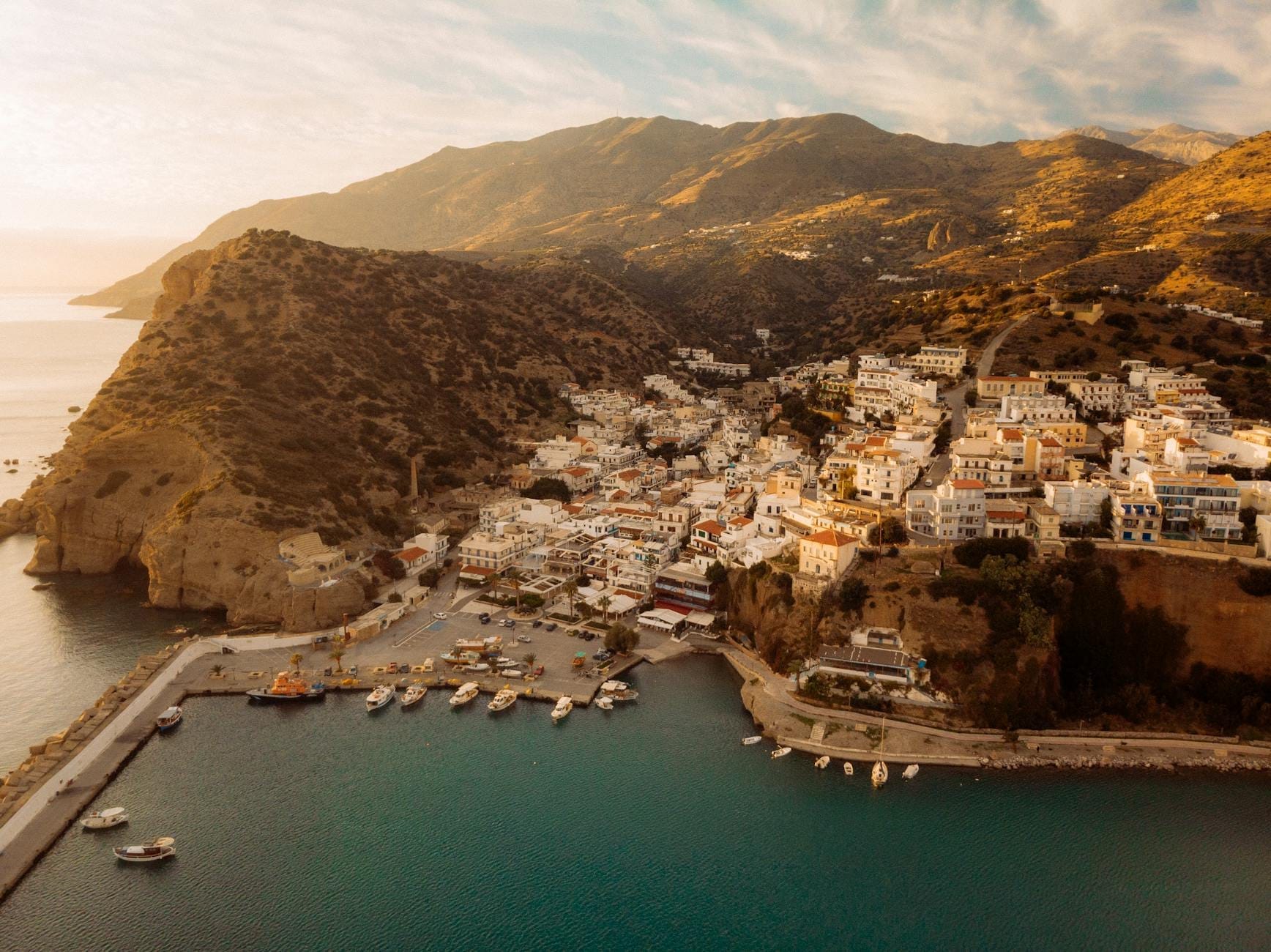Vista aerea di Agia Galini, un affascinante villaggio di pescatori con case bianche arroccate su ripide colline affacciate su un porto riparato con barche, incorniciato da montagne scenografiche durante l'ora d'oro.
