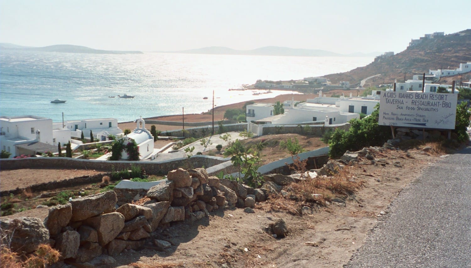 Vista de la playa Agios Ioannis en Mykonos, con edificios encalados, una orilla rocosa y mar turquesa bajo el brillante sol de la tarde.