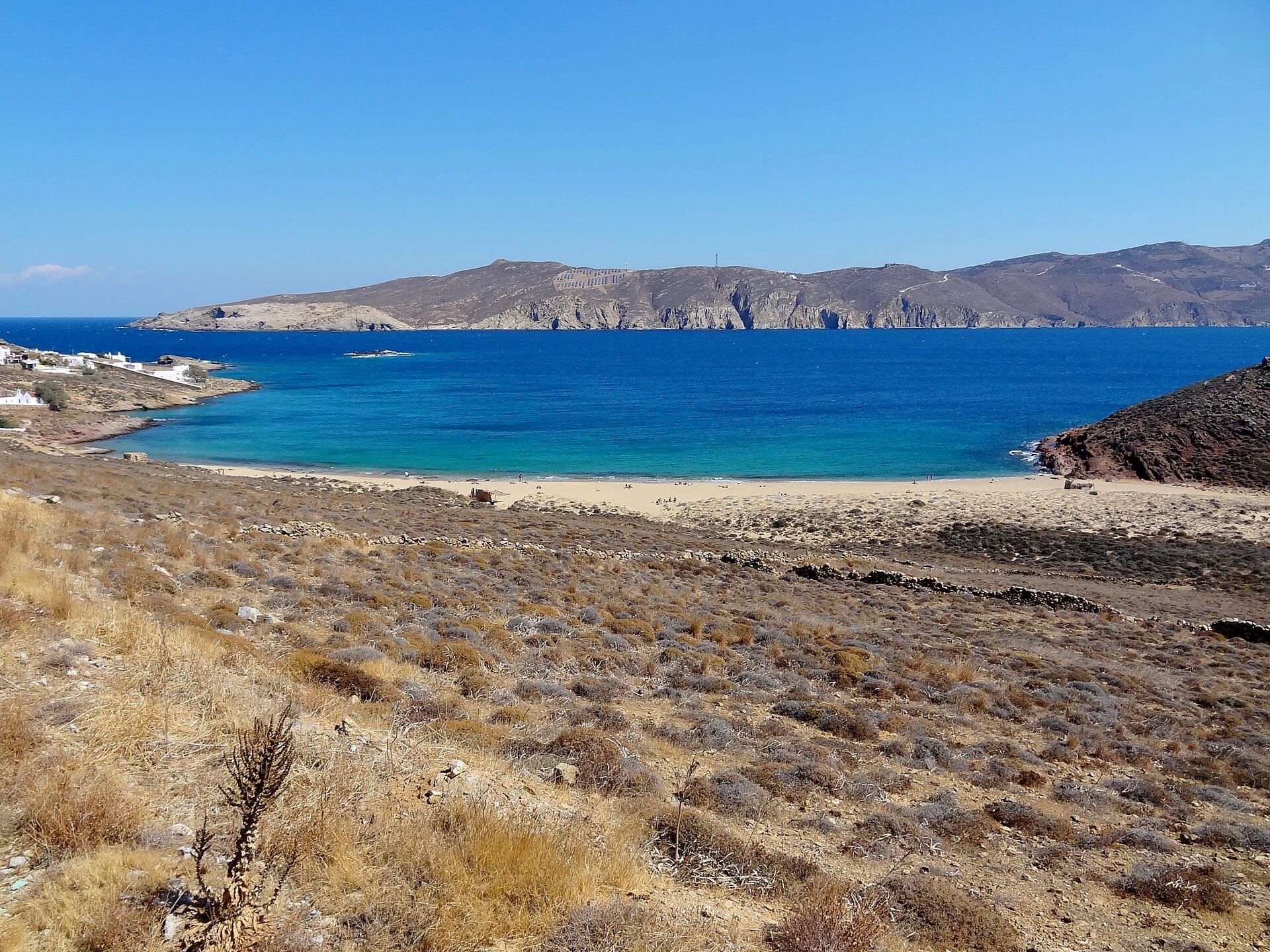 Vista amplia de la playa de Agios Sostis en Mykonos con arena dorada, mar azul cristalino, colinas áridas y edificios encalados dispersos.