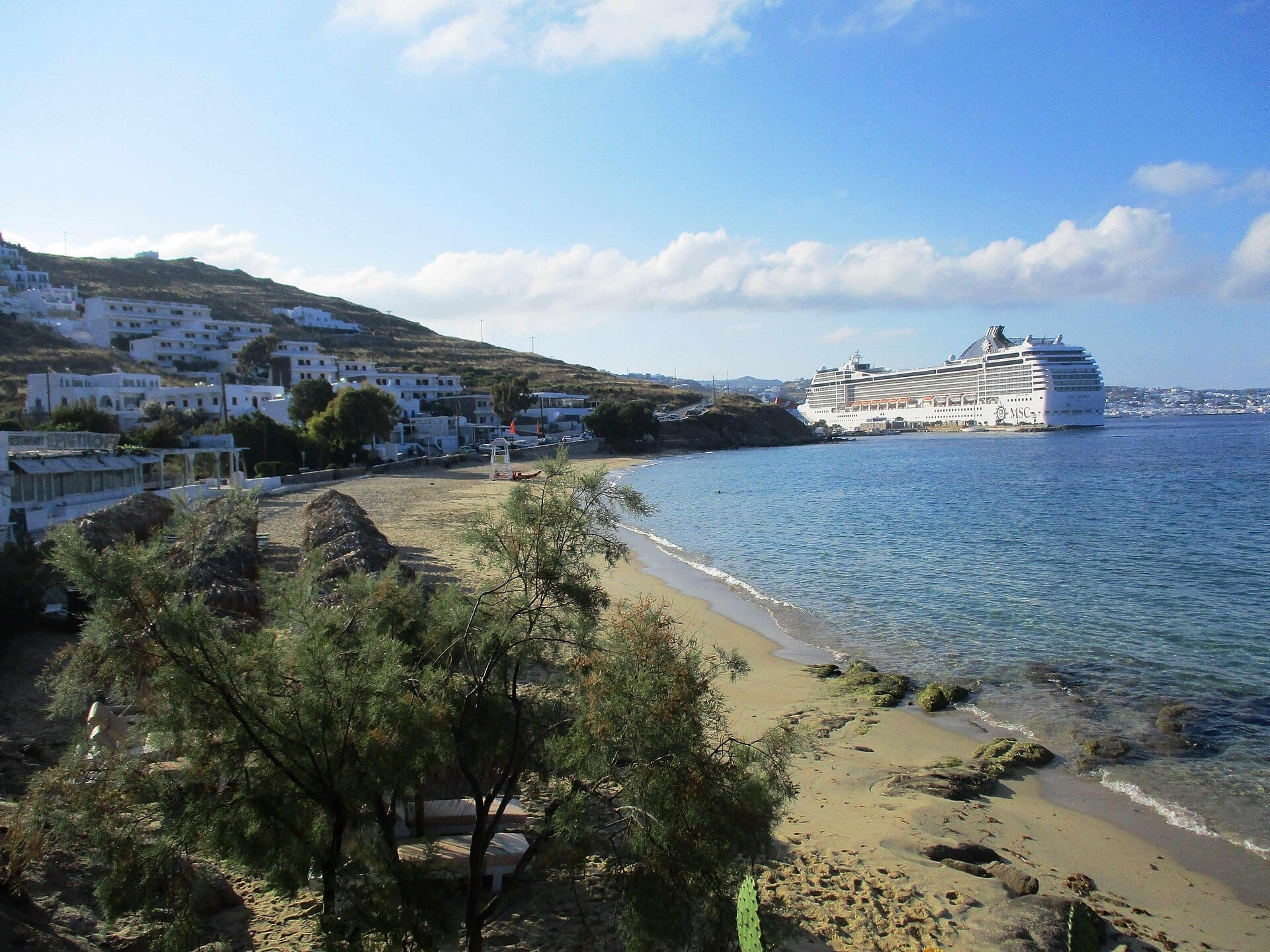 Playa de Agios Stefanos con una tranquila orilla de arena, edificios encalados en la ladera y un gran crucero atracado cerca de la costa bajo cielos azules.