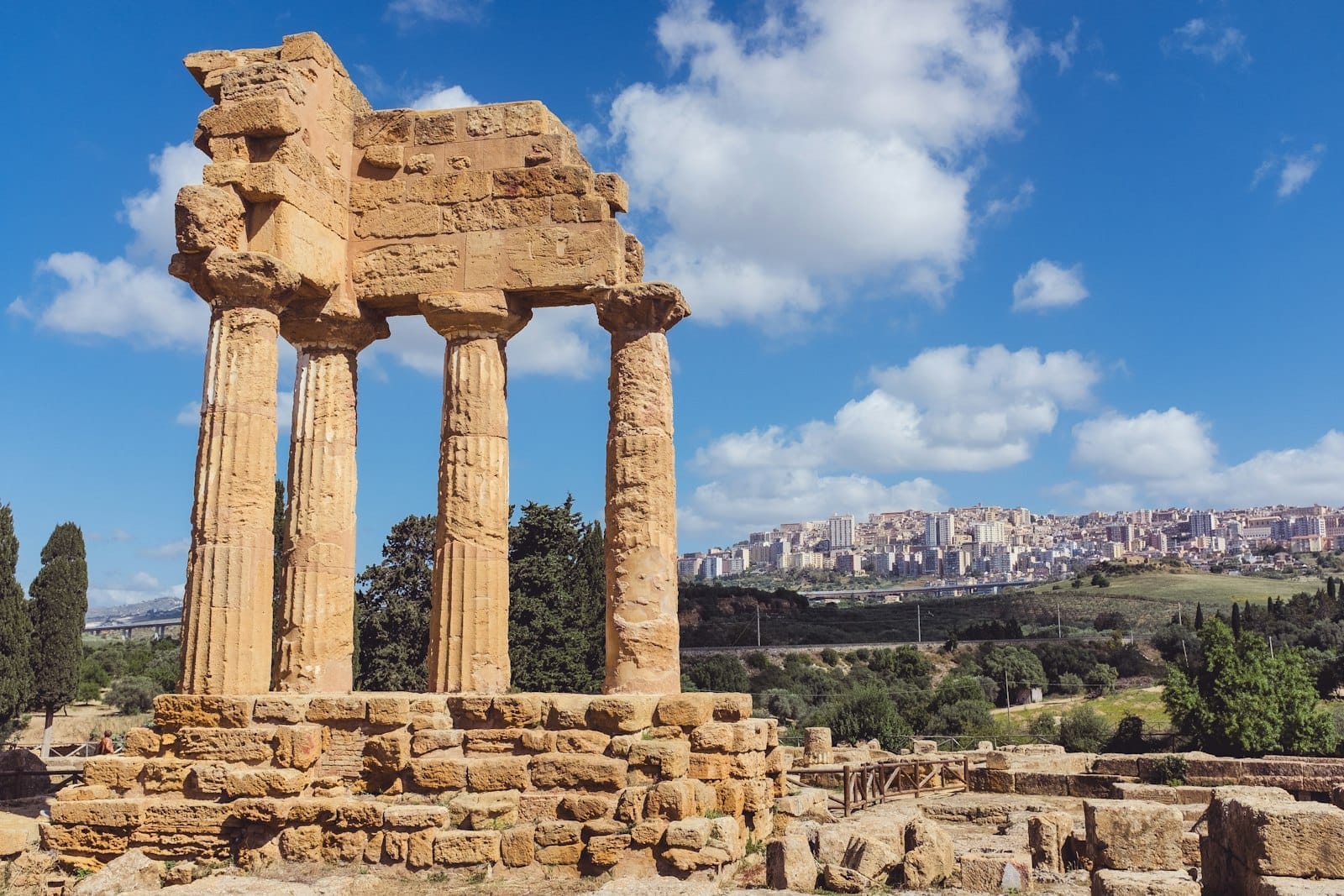 Antike griechische Tempelruinen im Tal der Tempel in Agrigento, Sizilien, mit Stadtsilhouette und grünen Hügeln im Hintergrund unter blauem Himmel.