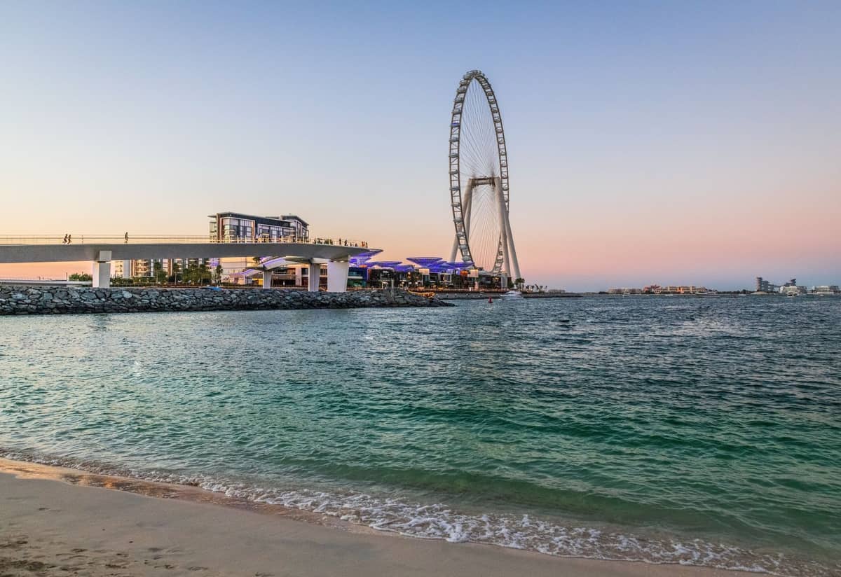 Weitwinkelpanorama des Ain-Dubai-Riesenrads, das bei Sonnenuntergang über der Bluewaters Island aufragt – im Vordergrund ruhiges Wasser und der Arabische Golf.
