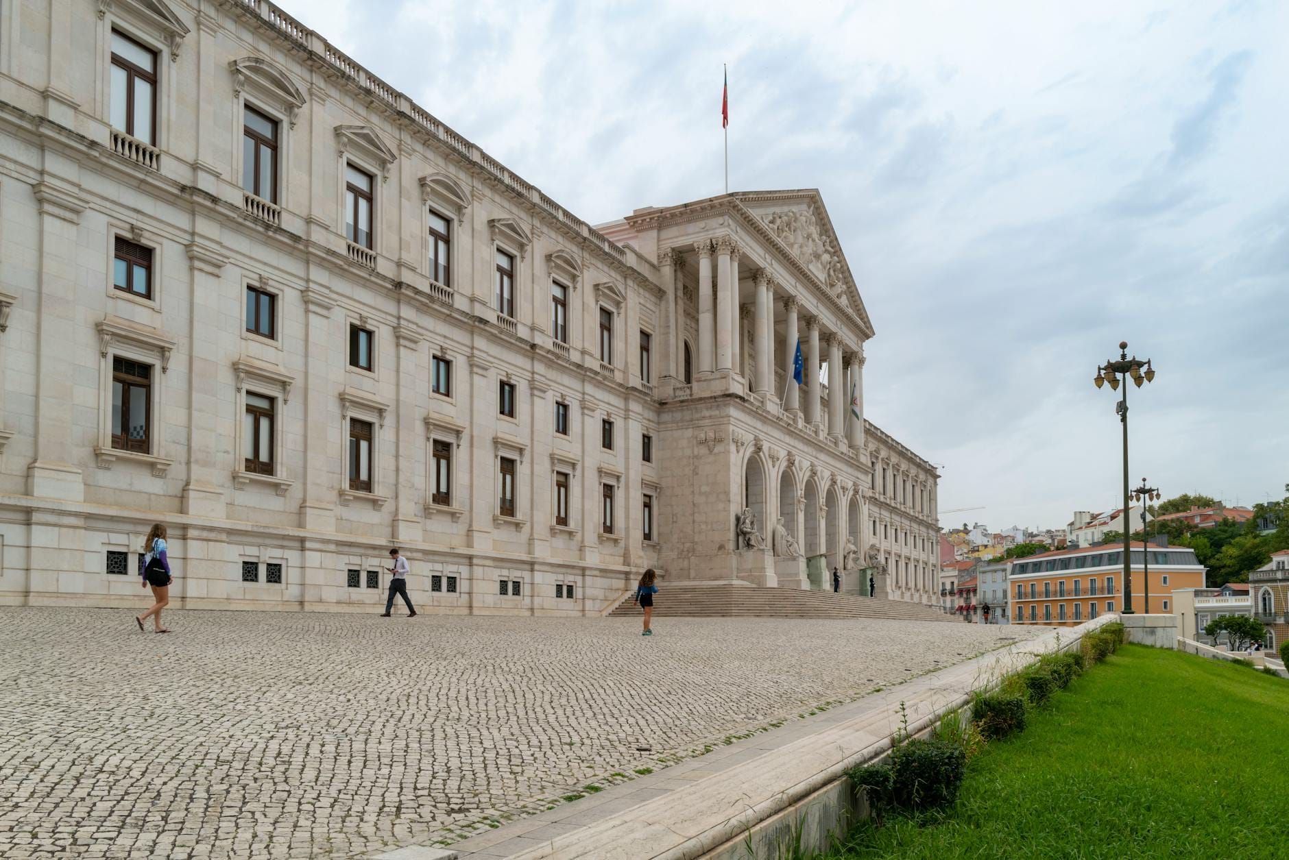 Vista grandangolare della facciata neoclassica del Palazzo Nazionale di Ajuda a Lisbona, con pedoni sulla piazza di sampietrini e un prato verde in primo piano sotto un cielo nuvoloso.