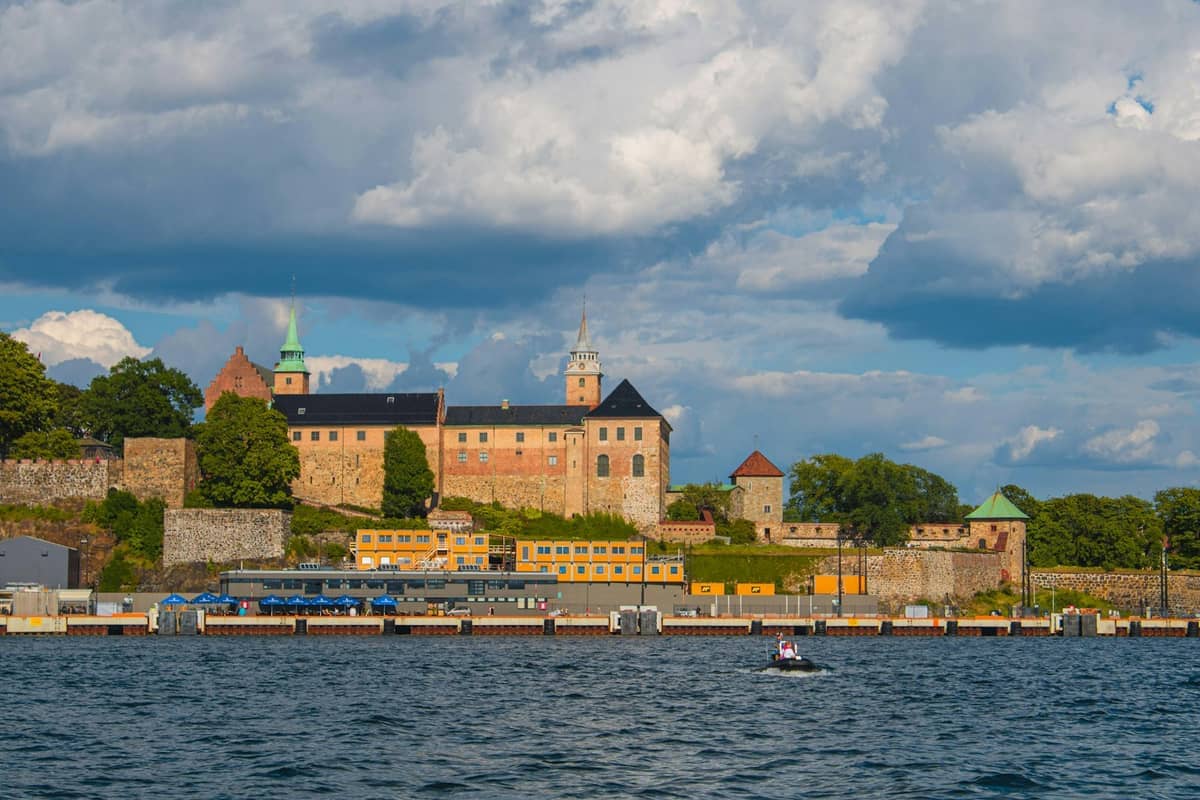 ป้อมปราการ Akershus พร้อมกำแพงหิน หอคอย และหลังคาสีเขียวที่มองเห็น Oslofjord ใต้เมฆก้อนใหญ่ในวันฤดูร้อน