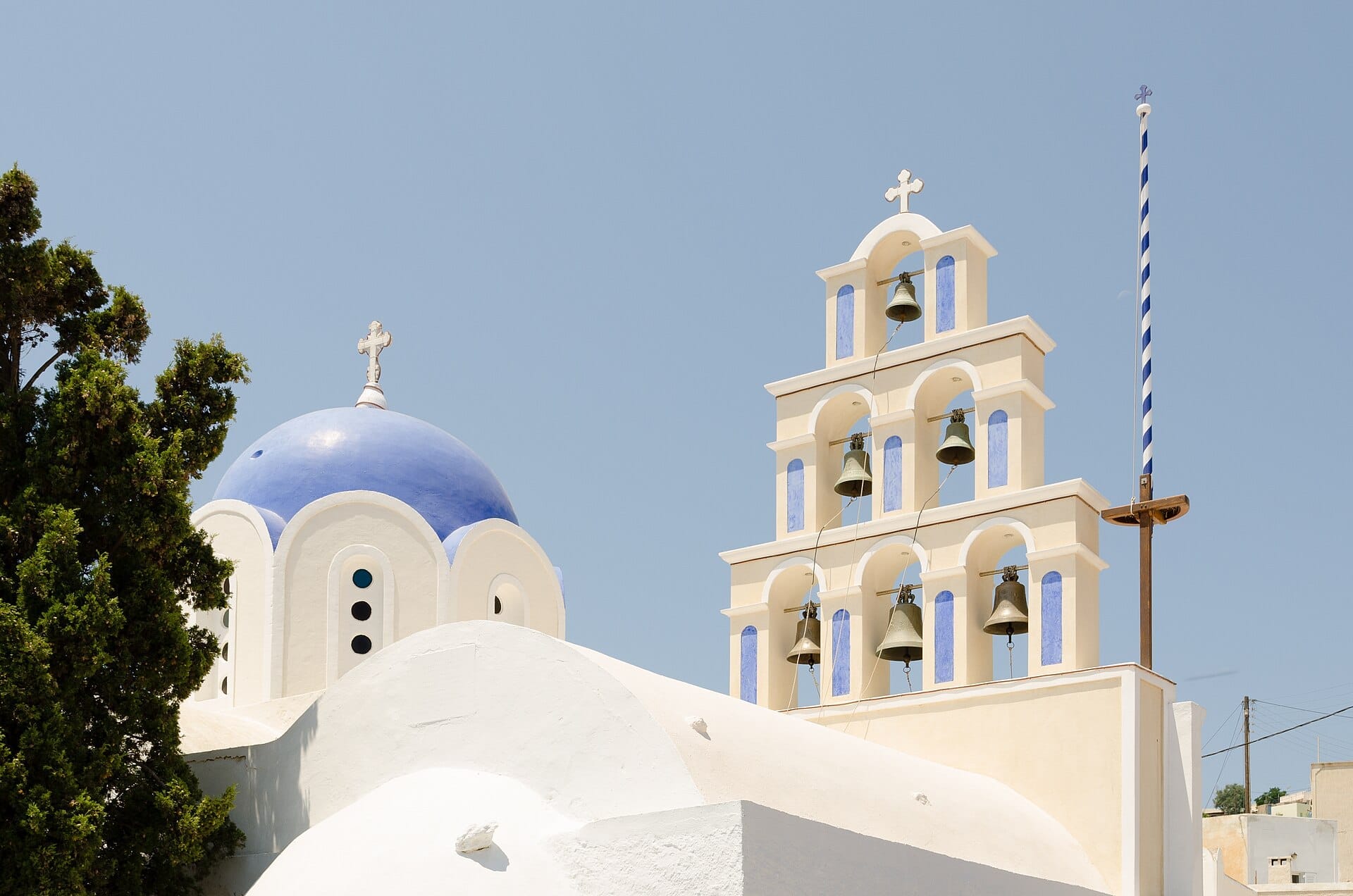 โบสถ์ทาสีขาวพร้อมโดมสีน้ำเงินและหอระฆังแบบดั้งเดิมใน Akrotiri, Santorini ใต้ท้องฟ้าสีฟ้าใสและแสงแดดจ้า