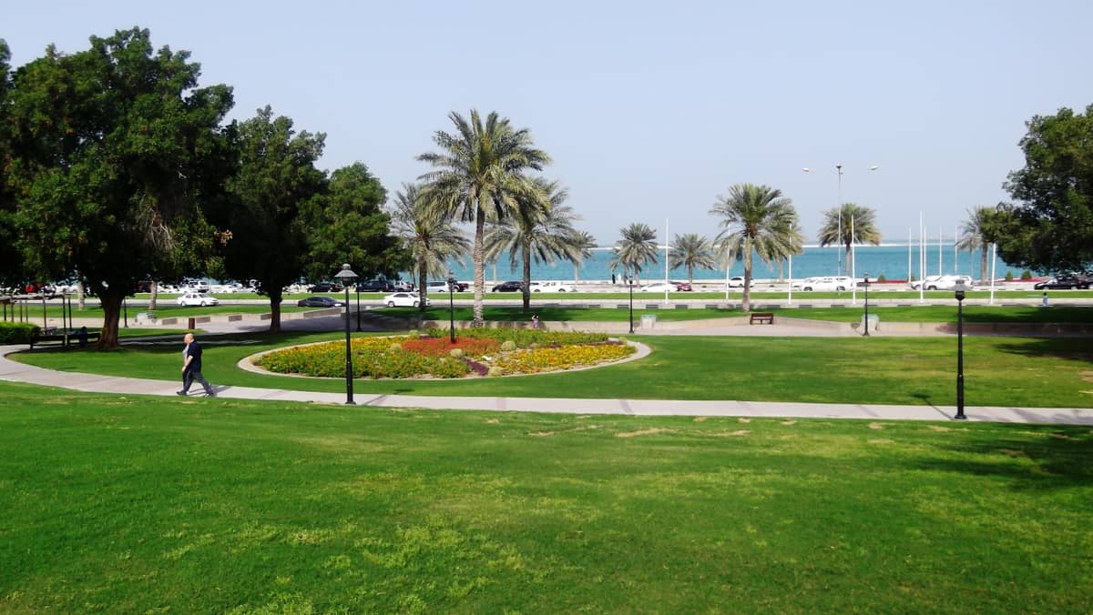 Weitläufige Parklandschaft des Al Bidda Parks mit grünen Rasenflächen, Palmen, Spazierwegen, Blumenbeeten und Blick auf die Corniche und das blaue Meer in Doha.