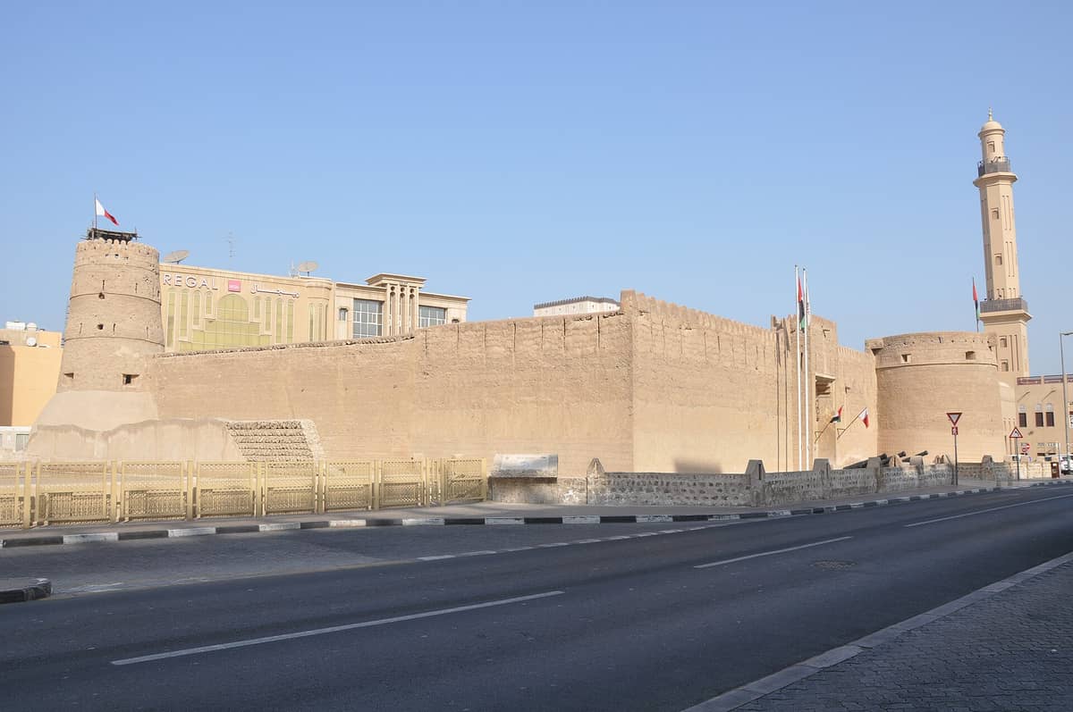 Weiter Blick auf das Al Fahidi Fort, Dubais ältestes Gebäude, mit seinen dicken Lehmziegelwänden, runden Türmen und emiratischen Flaggen unter einem strahlend blauen Himmel.