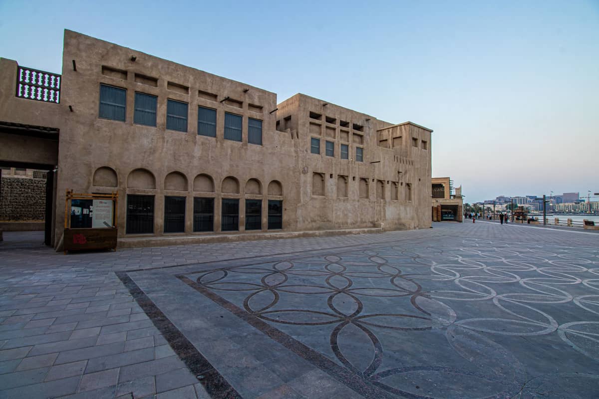 Ein weitläufiger, gepflasterter Platz, gesäumt von traditionellen sandfarbenen Gebäuden und Windtürmen im Al Fahidi Historischen Viertel in Dubai, in der Abenddämmerung.