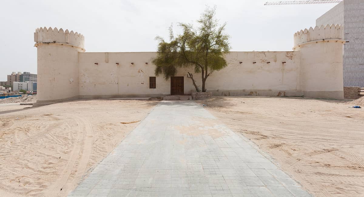 Frontale Ansicht des Al Koot Fort mit weiß getünchter Fassade, zwei runden Ecktürmen, zentralem Eingangstor und einem Baum, umgeben von sandigem Boden in Doha, Katar.