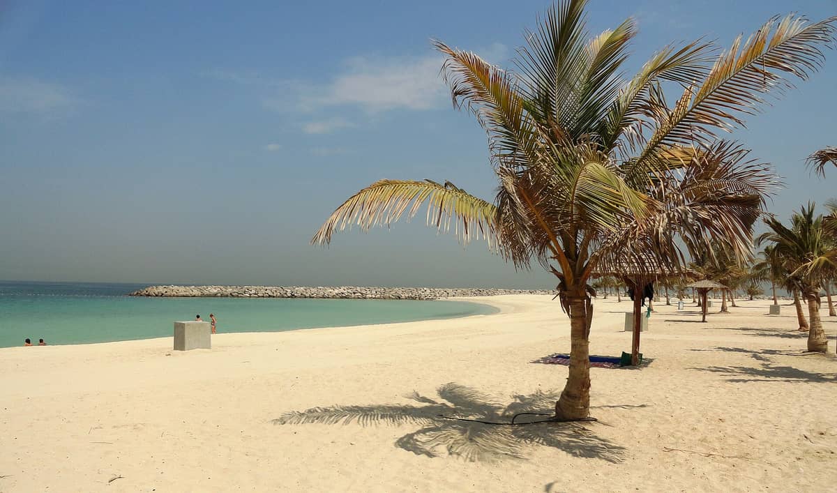Palmen und Sonnenschirme am weißen Sandstrand des Al Mamzar Beach Parks mit türkisfarbenem Wasser und einem Wellenbrecher unter strahlend blauem Himmel.