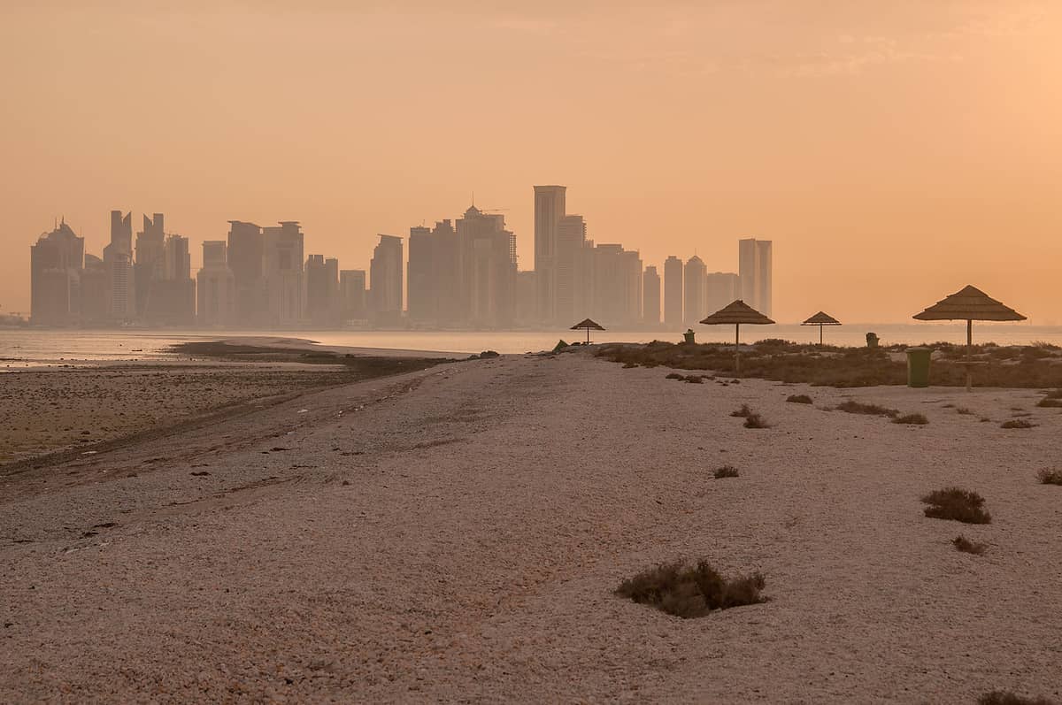 Verlassener Sandstrand auf der Insel Al Safliya mit vereinzelten Strohdächern und der Skyline von Doha im Hintergrund bei Sonnenuntergang.