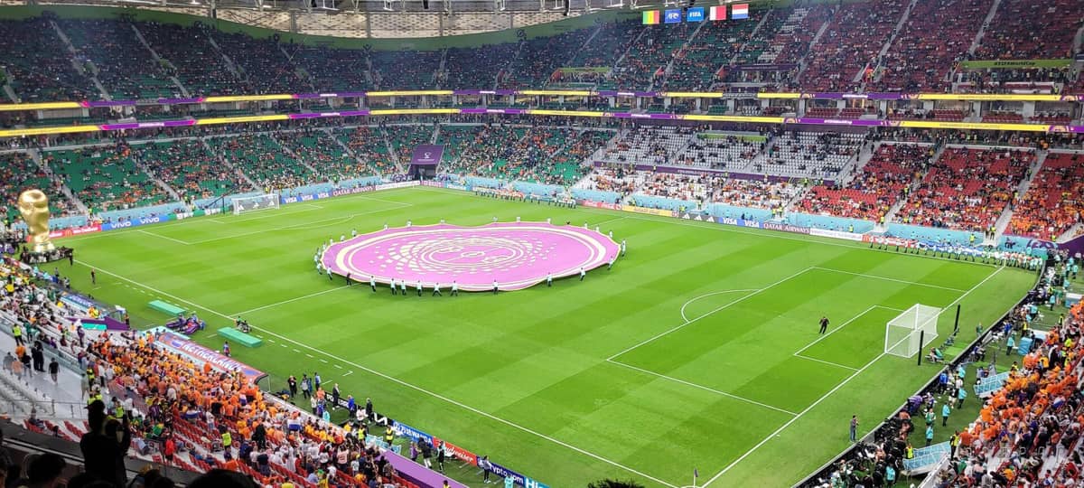 Weitwinkelaufnahme des Innenraums des Al Thumama Stadiums während eines FIFA-WM-Spiels: grüner Rasen, vollbesetzte Tribünen und ein großes WM-Display in der Spielfeldmitte.